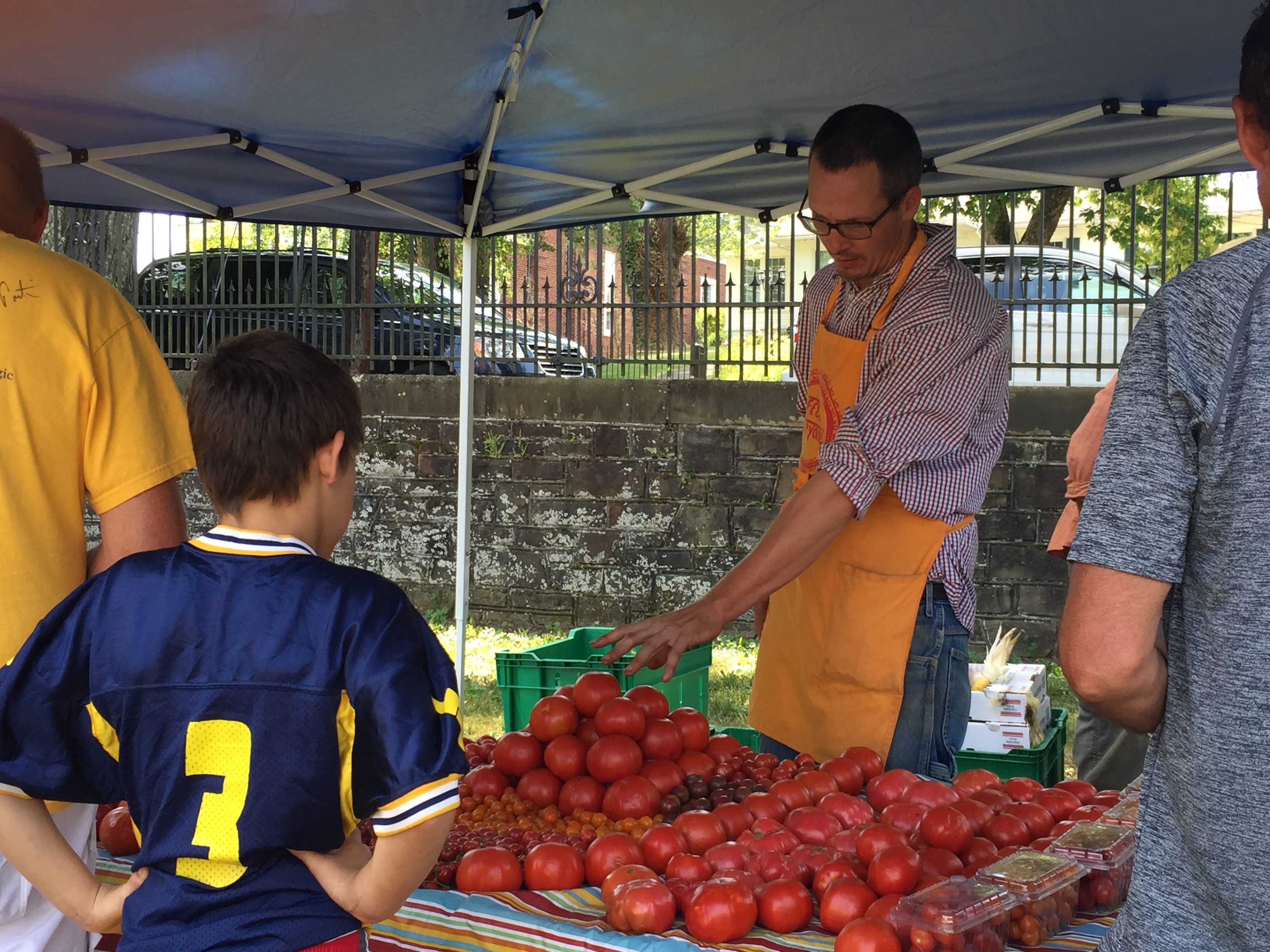 man standing behind a table with lots of red tomatoes talking to a young boy