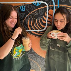 two girls standing in front of a coffee-themed mural sipping hot coffee and a cold coffee drink
