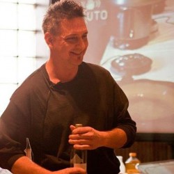 a man wearing a black top holding a bottle of wine while demonstrating a recipe during an Italian cooking class