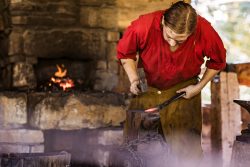 man dressed in a red shirt hammering in a blacksmith shop