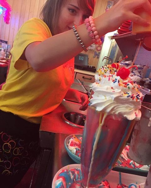 girl adding sprinkles to a milkshake that is topped with whipped cream and a cherry