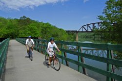 two people riding bicycles on a paved trail with a bridge in the background