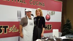 s woman dressed in black standing by a man wearing a hat and white apron as they demonstrate Italian family recipes during a cooking school.