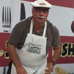 man wearing white apron and hat preparing food during a cooking demonstration