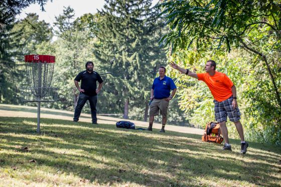 three guys in a wooded area throwing a disc into a metal basket