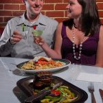 couple clinking cocktail glasses with plates of food on the table
