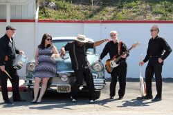 5 people dressed in black & white standing around a blue car