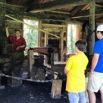 boy in yellow shirt and boy in blue shirt with hat watching a blacksmith demonstration
