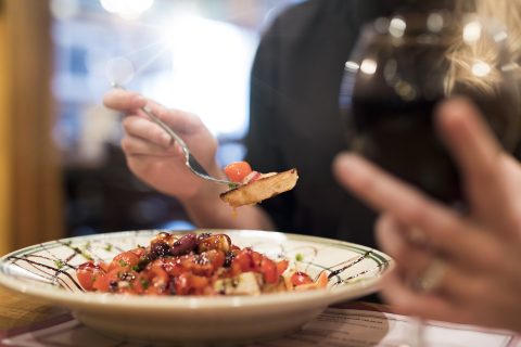 person holding a forkful of bruschetta and glass of red wine