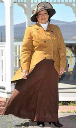 woman dressed in large hat with dark yellow jacket and long brown skirt