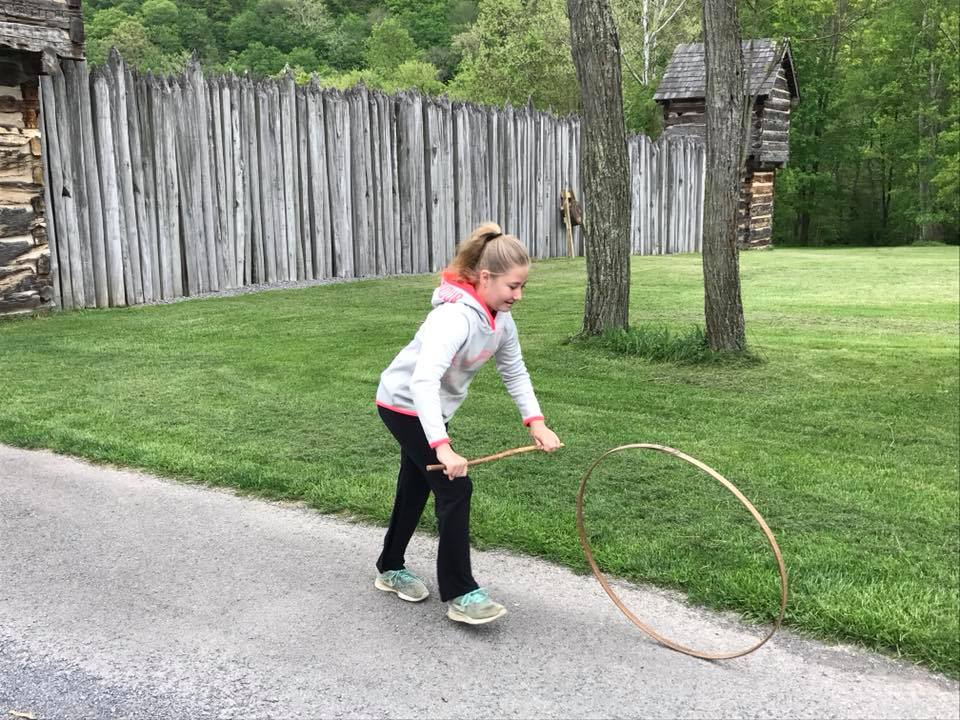 girl holding a stick trying to roll a wooden ring outside