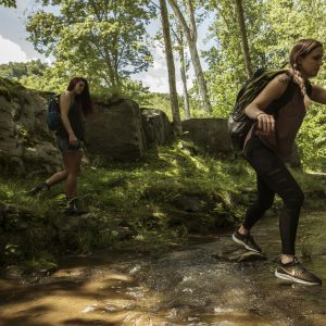 2 females jumping over a stream surrounded by rocks and trees
