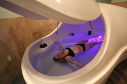 a woman in a black bikini floating in a large pod containing water with a purple light