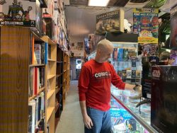 min in red shirt and jeans standing behind the glass display counter in a comic book store