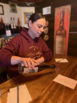 woman in burgundy sweatshirt standing at a counter pouring wine in a glass