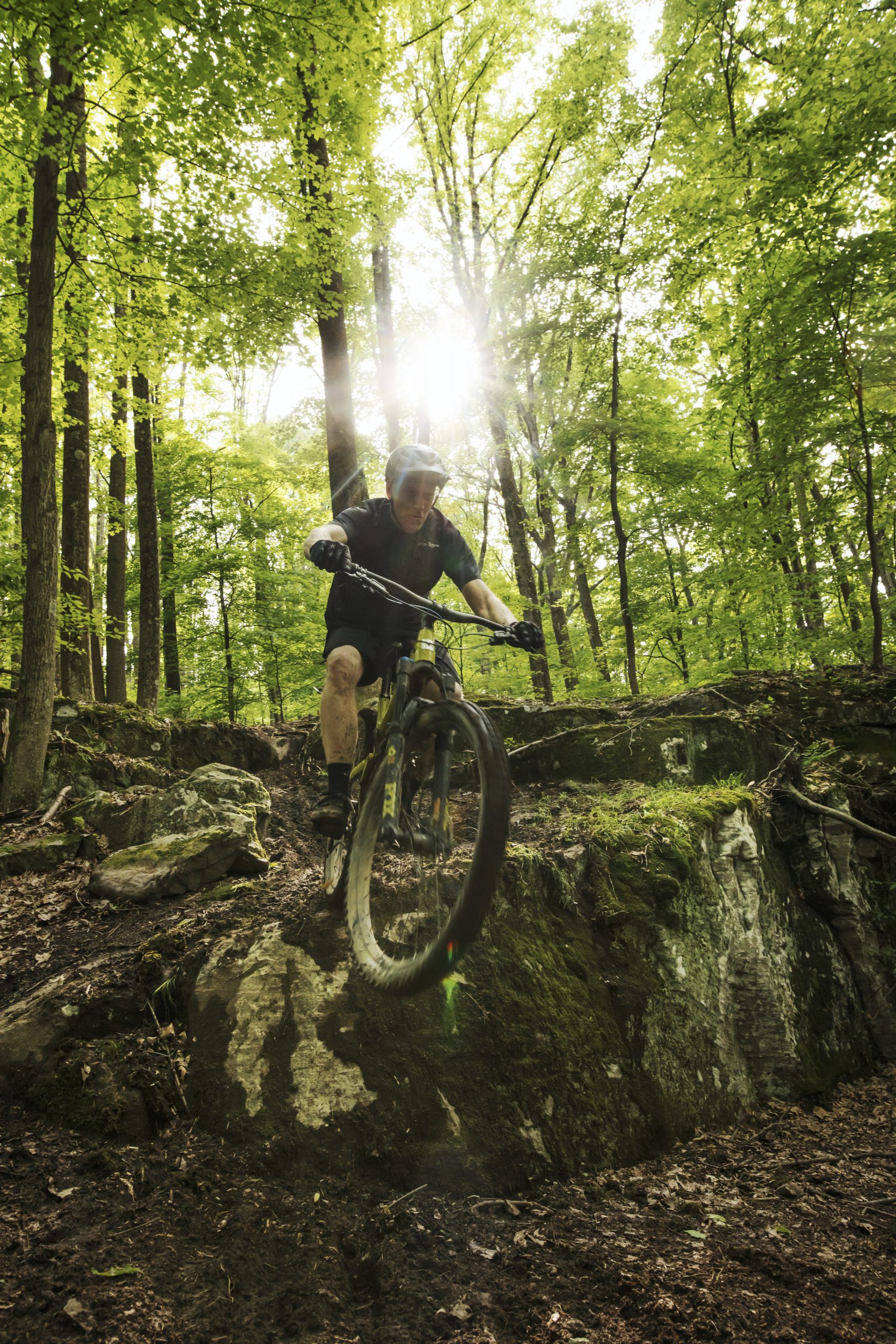 man riding a mountain bike over a large rock in the middle of the woods