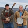 two men standing outside one is holding a banjo and the other a guitar