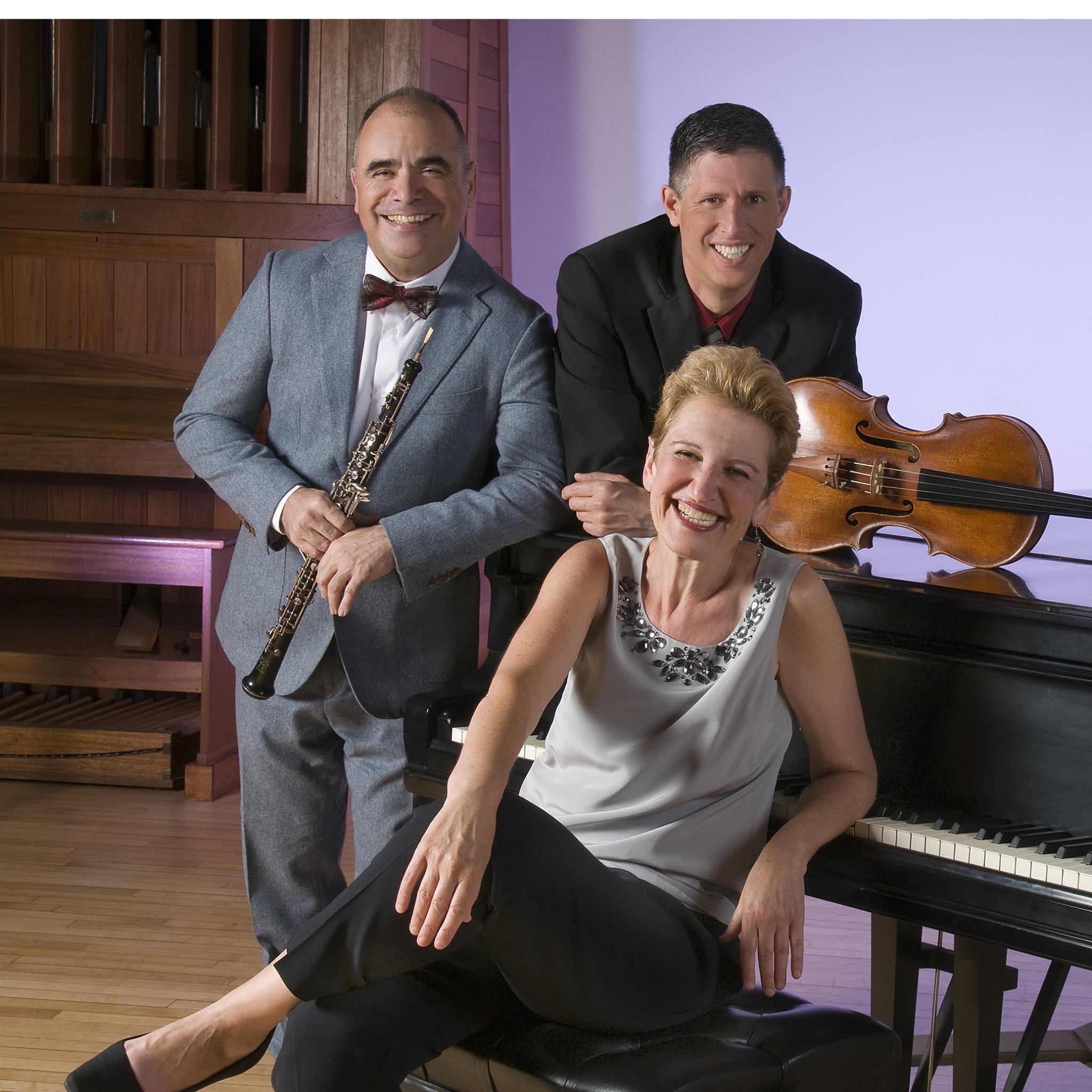 man in a gray suit standing holding an oboe, a man in a black suit leaning against a piano and a woman in a gray top sitting on a bench in front of a piano