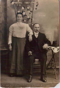 old sepia photo of a couple of a man sitting in a chair and a wife standing beside him