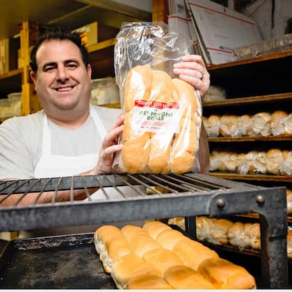 man standing behind a bakery counter holding a bag of pepperoni rolls