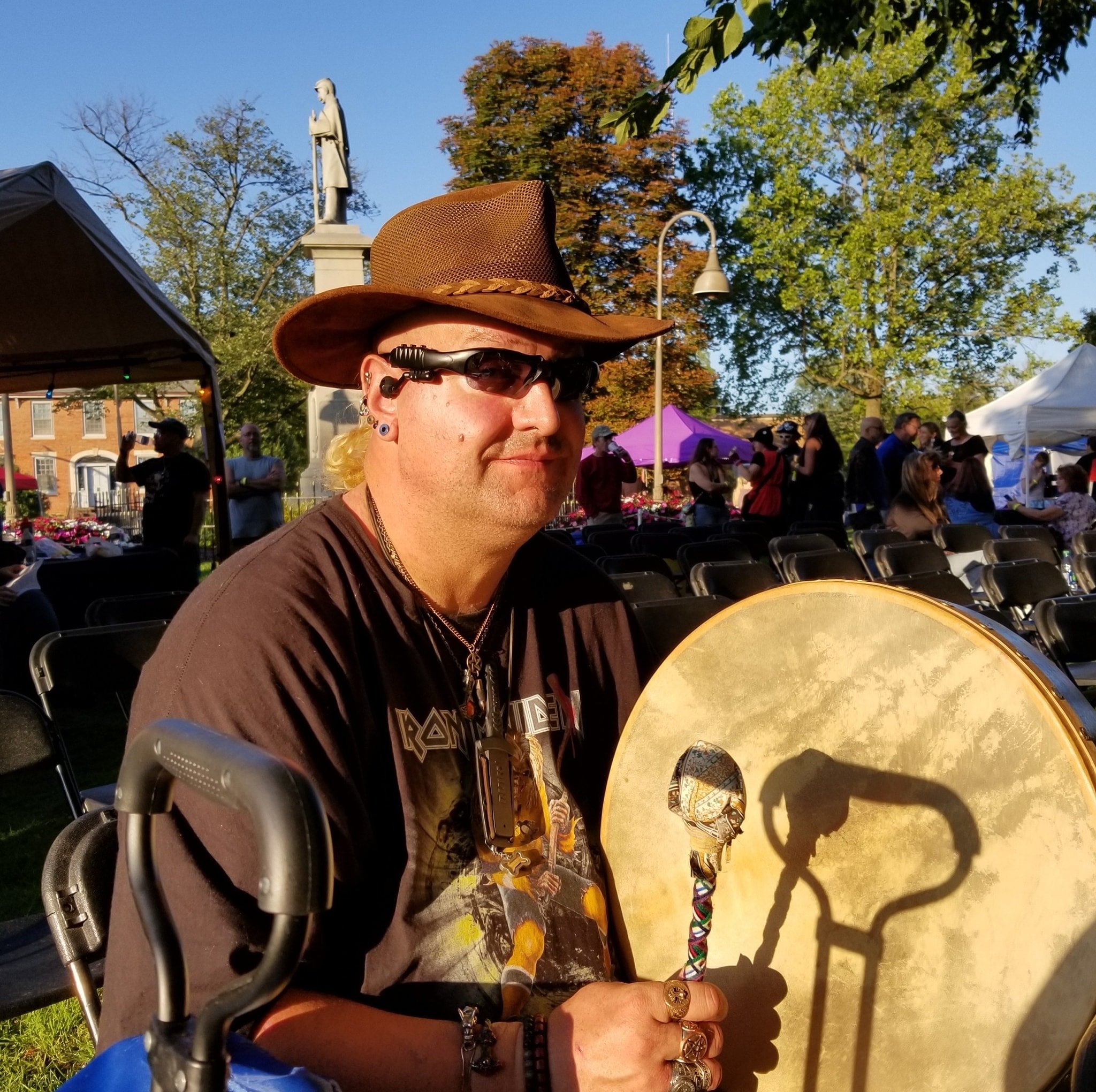 Man in brown hat and t-shirt wearing Native American jewelry holding a Native American drum stick and drum