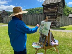 man in blue shirt and straw hat standing in front of a canvas on an easel painting a stockade fort