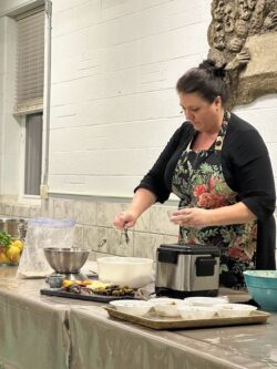 woman wearing a black top with floral apron demonstrating an Italian recipe during a cooking show.