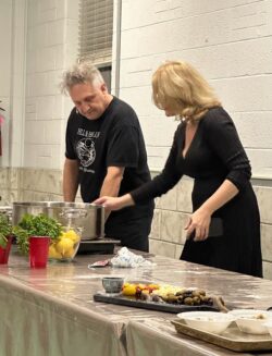 a man and a woman both dressed in black standing at a long table demonstrating how to make an Italian dish during a cooking class